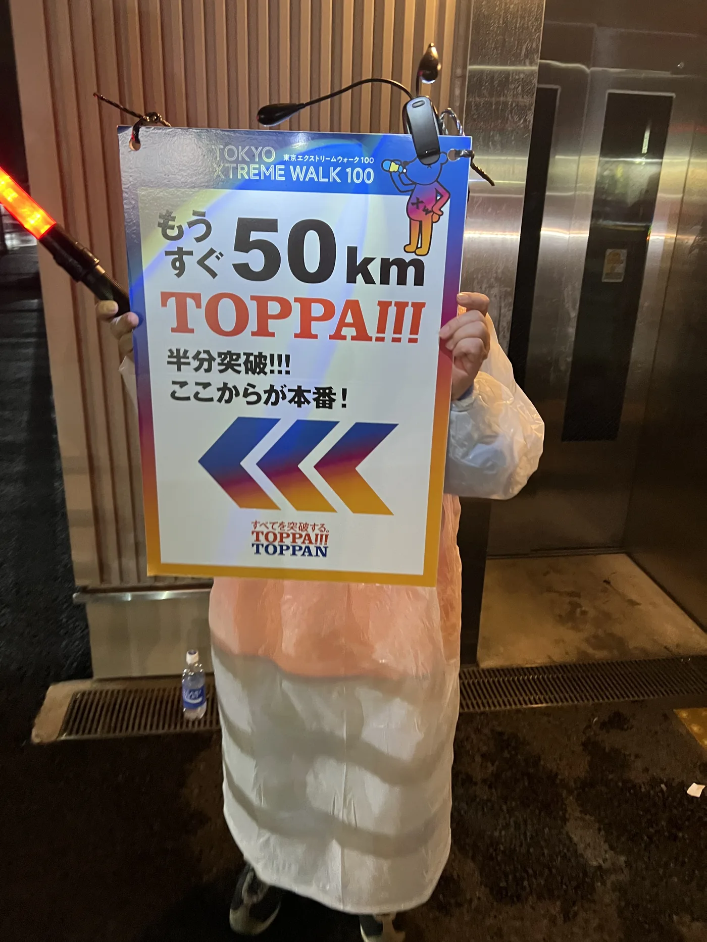 東京エクストリームウォーク100の「もうすぐ50km TOPPA!!!」の看板を持つ筆者。夜のチェックポイントでカッパを着ている