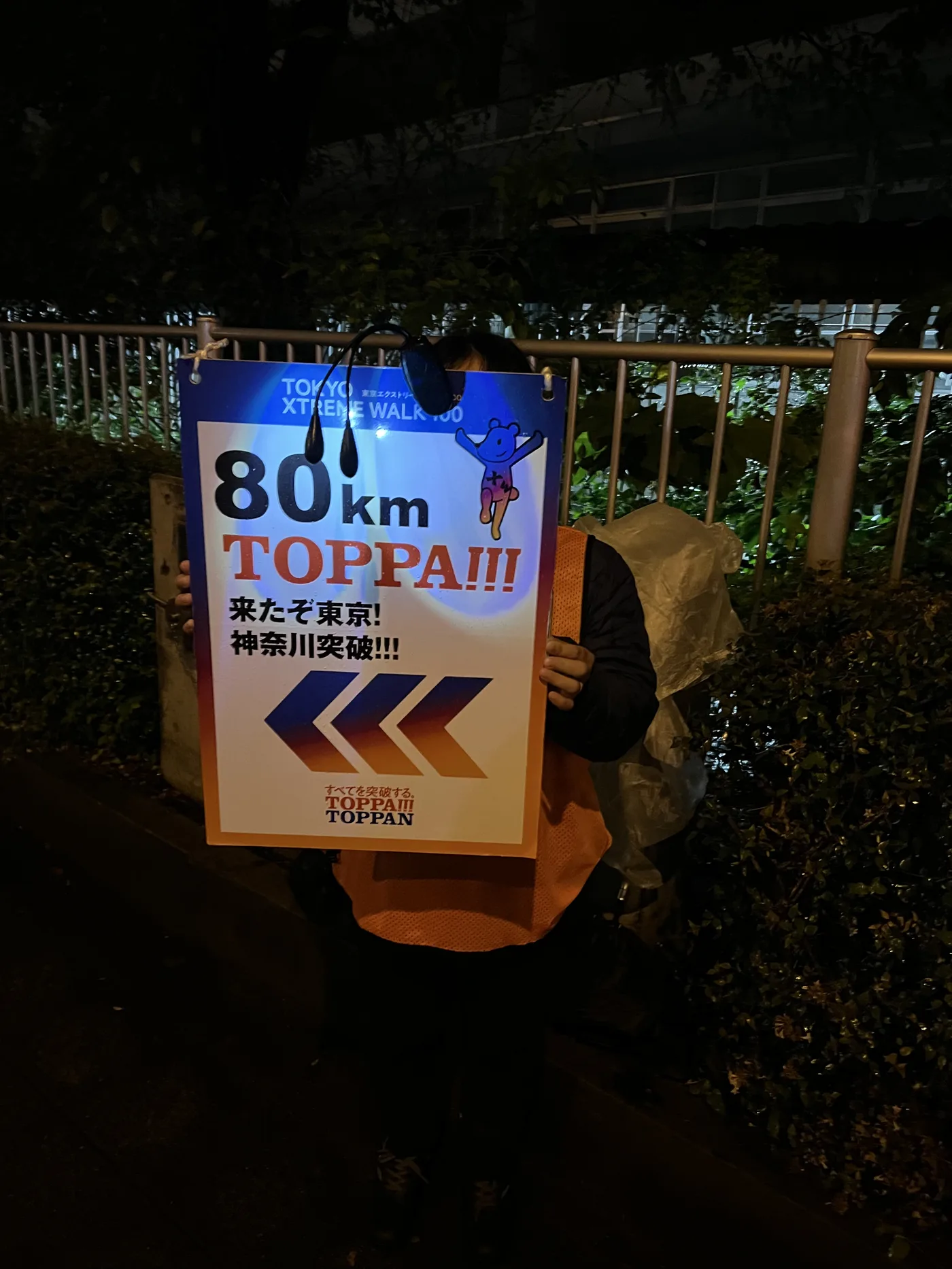 80km地点の看板「来たぞ東京！神奈川突破!!!」を持つ筆者。リュックにビニール袋をかぶせている